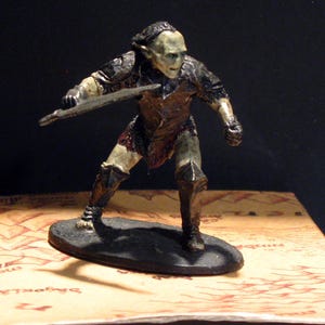 May include: A painted miniature figurine of an Orc warrior, wearing armour and holding a sword. The Orc has a green face and is standing on a black base.