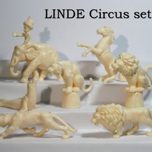 Puede incluir: Colección de figuras de animales de circo de color blanquecino, que incluyen un elefante con una figura encima, un caballo encabritado, leones, un tigre y una foca. El texto "LINDE Circus set B" se muestra encima.