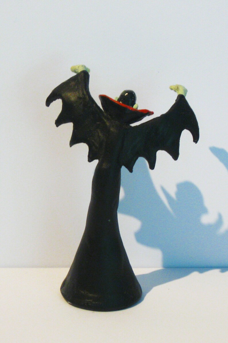 May include: A black and red plastic figurine of a vampire with outstretched wings. The vampire is wearing a black cape and a red hat.