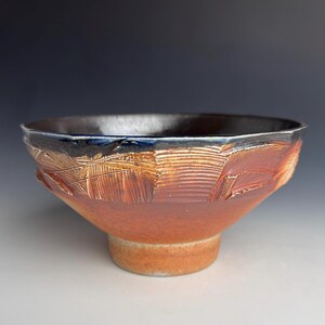 May include: A ceramic bowl with a wide, flared rim and a circular base. The exterior features a textured, reddish-brown finish with carved, linear patterns. The interior is glazed in a deep, dark black with a glossy sheen along the upper edge.