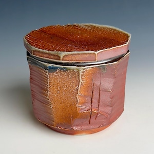 May include: A cylindrical ceramic jar with a matching lid, featuring a textured, multi-toned surface in shades of burnt orange, deep red, and brown. The exterior displays vertical ridges, subtle indentations, and a mottled, rustic glaze finish.