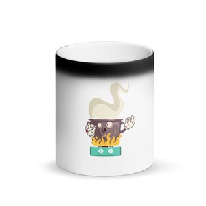 Könnte beinhalten: Weißer Keramikbecher mit schwarzem Oberteil. Der Becher zeigt eine Cartoon-Illustration eines Kochtopfs mit Flammen, Dampf und einem überraschten Gesichtsausdruck. Der Topf steht auf einem grünen Herd, wobei der Topf eine „Okay“-Handgeste macht.