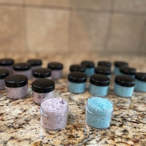 May include: A close-up of two small glass jars filled with bath salts. The jar on the left contains purple bath salts, and the jar on the right contains blue bath salts.