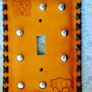 May include: A rectangular, orange leather light switch cover with a black braided border. The cover features silver-colored rivets, a single switch opening, and etched images of a buffalo and a bull skull with feathers.