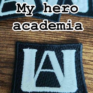 Może przedstawiać: Czarne, haftowane naszywki z napisem "My hero academia" i literami "UA" w kolorze białym. Prostokątne naszywki mają czarną obw&oacute;dkę i są umieszczone na drewnianej powierzchni.