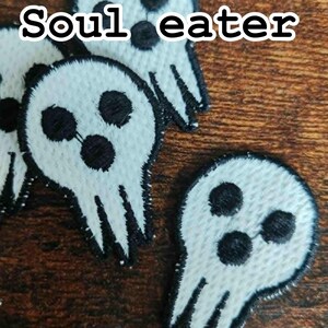 Może przedstawiać: Białe, haftowane naszywki z motywem czaszki i czarnym napisem "Soul eater". Naszywki mają trzy czarne kropki dla oczu i usta, z czarnym obrysem. Na brązowej drewnianej powierzchni.
