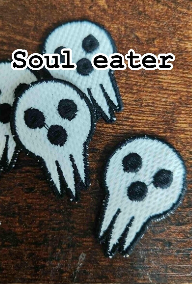 Może przedstawiać: Białe, haftowane naszywki z motywem czaszki i czarnym napisem "Soul eater". Naszywki mają trzy czarne kropki dla oczu i usta, z czarnym obrysem. Na brązowej drewnianej powierzchni.