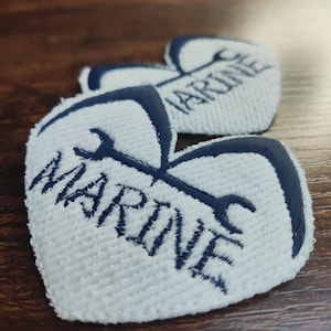 Peut inclure: Trois écussons en forme de cœur avec le mot "MARINE" brodé en bleu marine sur un fond blanc texturé. Un motif de clé à molette bleu marine est au-dessus du mot. Les écussons sont disposés sur une surface en bois.
