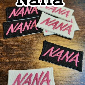 Może przedstawiać: Haftowane naszywki z napisem "Nana" w r&oacute;żowym skrypcie. Niekt&oacute;re naszywki są na czarnym materiale, inne na białym. Naszywki są prostokątne i mają obszyte obramowanie. Słowo "Nana" jest r&oacute;wnież wyświetlane powyżej.