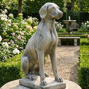 Könnte beinhalten: Eine verwitterte Hundestatue aus Stein sitzt auf einem runden Sockel in einem Garten. Der Hund sitzt, der Kopf zur Seite gedreht. Im Hintergrund sind Bäume, Büsche und ein Steinweg zu sehen. Dekorative Gartenverzierung.
