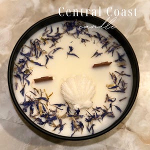 May include: A round black candle tin with a creamy white wax. The candle is decorated with blue flower petals, a white seashell, and two small wooden sticks. The text "Central Coast candle" is written in white script.