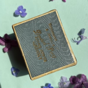 May include: A small, rectangular box with a textured, grey surface and gold lettering. The box is surrounded by small purple and blue flowers on a light green background. The box has gold trim.