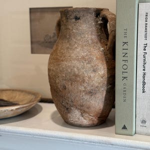 Könnte beinhalten: Eine verwitterte, braune Keramikvase mit zwei Henkeln. Die Vase wird mit zwei Büchern, "The Kinfolk Garden" und "The Furniture Handbook" von Frida Ramstedt, präsentiert. Eine Holzschale und ein gerahmtes Kunstwerk sind ebenfalls zu sehen.