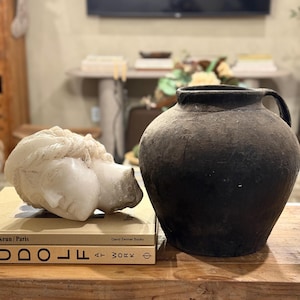Könnte beinhalten: Ein Stillleben mit einer weißen Skulptur eines Kopfes, die auf einem Stapel Bücher ruht. Rechts befindet sich eine große, dunkle, strukturierte Vase mit Henkel. Die Gegenstände stehen auf einer Holzoberfläche.