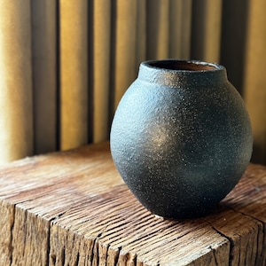 Puede incluir: Un jarrón de cerámica gris oscuro con una superficie texturizada se asienta sobre un bloque de madera rústico.
