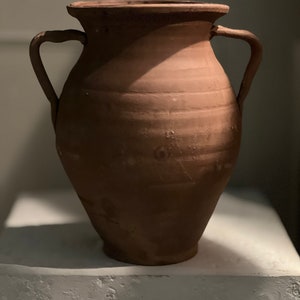 以下が含まれることがあります： 2つの取っ手のあるテラコッタ製の壺。壺は茶色で、表面がざらざらしている。