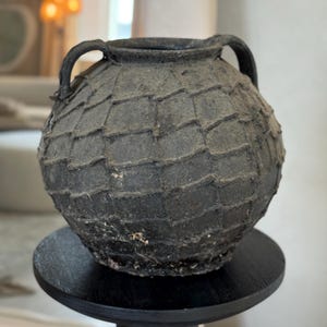 May include: A dark gray, textured, spherical vase with two handles. The vase has a scale-like pattern across its surface and sits on a small, round, black stand. The background is blurred, suggesting an indoor setting.