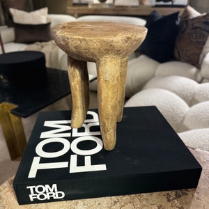 May include: A wooden stool with three legs. The stool is light brown and has a natural finish. The stool is sitting on top of a black book with the words "TOM FORD" printed in white.