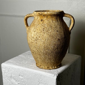 Könnte beinhalten: Eine rustikale, beige Keramikvase mit zwei Henkeln. Die Vase hat eine strukturierte Oberfläche und eine leicht abgerundete Form.