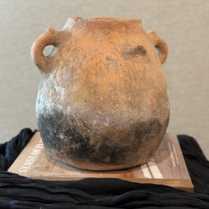 Puede incluir: Un jarrón de cerámica de color terracota desgastado con dos asas de bucle. El jarrón tiene un cuerpo redondeado y un borde ligeramente ensanchado. La parte inferior del jarrón muestra decoloración oscura. El jarrón está sobre un libro.