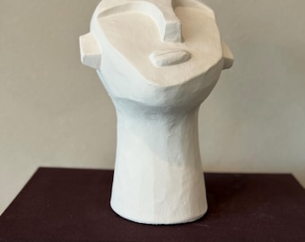 Könnte beinhalten: Eine weiße, abstrakte Skulptur eines menschlichen Kopfes mit vereinfachten Merkmalen. Der Kopf hat einen langen Hals und steht auf einer dunklen Oberfläche. Die Skulptur besteht aus einem strukturierten Material.