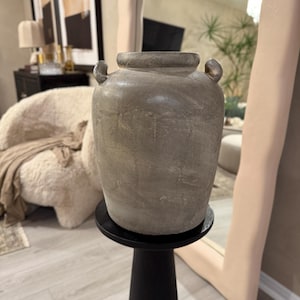 May include: A gray ceramic vase with two handles sits on a black pedestal. The vase has a textured surface and a slightly rounded shape.