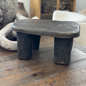 May include: A dark brown wooden stool with four legs. The stool is made from a single piece of wood and has a rounded top.