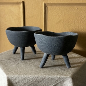 May include: Two black ceramic plant pots with three legs each. The pots are sitting on a light gray textured surface.