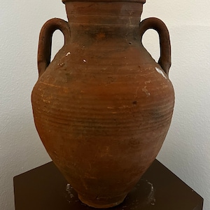 May include: A terracotta clay amphora with two handles, a rounded body, and a wide mouth. The amphora is sitting on a dark brown pedestal.