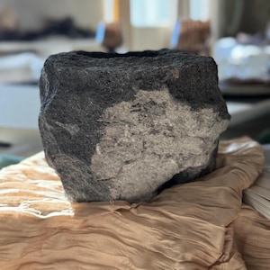 May include: A dark gray, rough-hewn stone vessel with a rectangular shape. The top opening is a dark gray, while the lower portion is a lighter gray. The vessel is resting on a tan fabric.