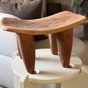 May include: A wooden stool with a curved seat and three legs. The stool is made of light brown wood with darker brown accents, showcasing the natural grain. The legs are tapered and provide a stable base. This unique piece adds a touch of rustic charm. The stool is approximately 30 cm high.