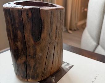 May include: A rustic, cylindrical wooden vase with a hollowed-out top. The vase is made from a section of a tree trunk, showcasing the natural grain and texture of the wood. The color is a warm brown with darker streaks, adding to its organic appeal.
