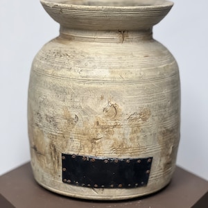 Puede incluir: Un jarrón de madera blanco con una placa de metal negro unida al frente. La placa tiene una fila de pequeños remaches de metal alrededor del borde.