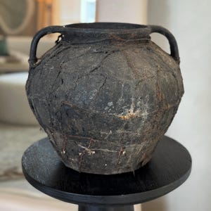 May include: A dark gray ceramic pot with two handles and a rough, textured surface. The pot is decorated with woven strips of material.