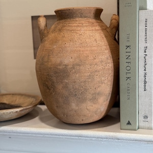 May include: A large, aged terracotta vase with two handles. The vase has a textured surface and a wide, rounded body. Beside the vase are two books, one titled "The Kinfolk Garden". The vase is a decorative item.