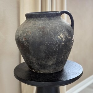 May include: A dark gray, rustic, ceramic pitcher with a handle. The pitcher has a worn, aged appearance and is sitting on a black pedestal.