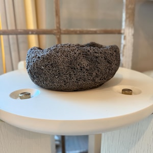 May include: A dark gray, textured stone bowl with an irregular rim sits on a white, round table. The table has two brass-colored accents. The bowl's porous surface contrasts with the smooth table, creating a natural, rustic aesthetic.