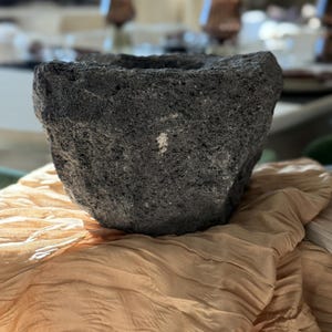 May include: A dark grey, rough-textured mortar and pestle. The bowl-shaped mortar has a wide opening and sits on a tan, pleated fabric. The mortar is made of a dark, volcanic rock.