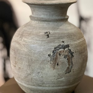 Puede incluir: Un jarrón de cerámica gris con cuerpo redondeado y un borde ancho y plano. El jarrón tiene un aspecto rústico y desgastado con líneas visibles y una mancha marrón en la parte delantera.