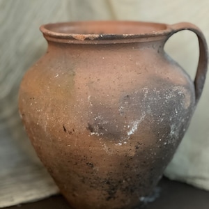以下が含まれることがあります： ハンドルが付いた茶色の粘土製のピッチャー。ピッチャーは、濃い茶色の斑点と白い筋のある、すり切れた素朴な外観をしています。