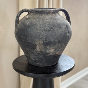 May include: A dark gray ceramic vase with two handles, sitting on a black pedestal. The vase has a rustic, aged appearance.