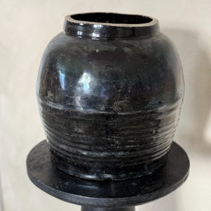 Puede incluir: Un tarro de cerámica negro con cuerpo redondeado y borde ligeramente acampanado. El tarro tiene un acabado brillante y está decorado con líneas horizontales.