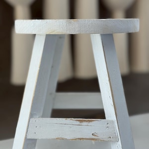Puede incluir: Un pequeño taburete de madera pintado de blanco, con asiento redondo y patas anguladas. El taburete tiene un acabado envejecido, con vetas de madera visibles y ligeras imperfecciones. El diseño es sencillo y rústico.