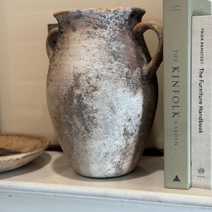 Puede incluir: Un jarrón de cerámica texturizado con dos asas, en tonos blancos, grises y marrones. El libro The Kinfolk Garden y The Furniture Handbook son visibles en el fondo. Es un objeto decorativo.