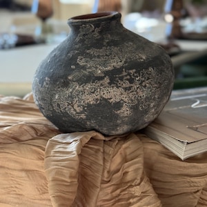 May include: A dark grey, textured ceramic vase with a rounded body and a narrow opening. The vase has a weathered appearance with lighter patches, sitting on a beige, draped fabric. The vase is a decorative home decor item.