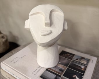 Könnte beinhalten: Eine weiße, minimalistische Skulptur eines menschlichen Kopfes mit vereinfachten Merkmalen, darunter Nase, geschlossene Augen und Mund. Die Skulptur steht auf einem Stapel Bücher mit den Worten "Soft Minimal".