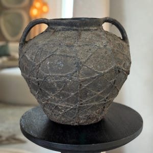 May include: A dark gray ceramic vase with two handles and a textured, geometric pattern. The vase is sitting on a black pedestal.