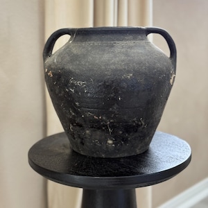 May include: A black ceramic pot with two handles. The pot has a rounded body and a slightly flared lip. It is sitting on a black pedestal.