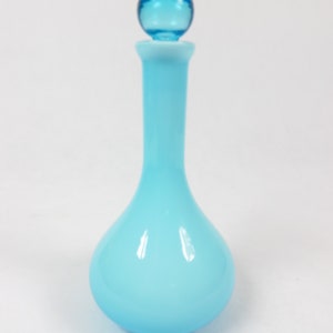 Könnte beinhalten: Eine hellblaue Glasflasche mit einem runden, hellblauen Glasstopfen. Die Flasche hat einen abgerundeten Boden und einen langen, dünnen Hals.