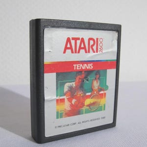 Pode incluir: Um cartucho de videogame Atari 2600 preto para o jogo Tennis. O rótulo apresenta o logotipo vermelho da Atari 2600, a palavra Tennis em branco em uma barra vermelha e uma ilustração de dois tenistas em uma quadra. O texto 1987 ATARI CORP. ALL RIGHTS RESERVED 2680 está impresso na parte inferior.